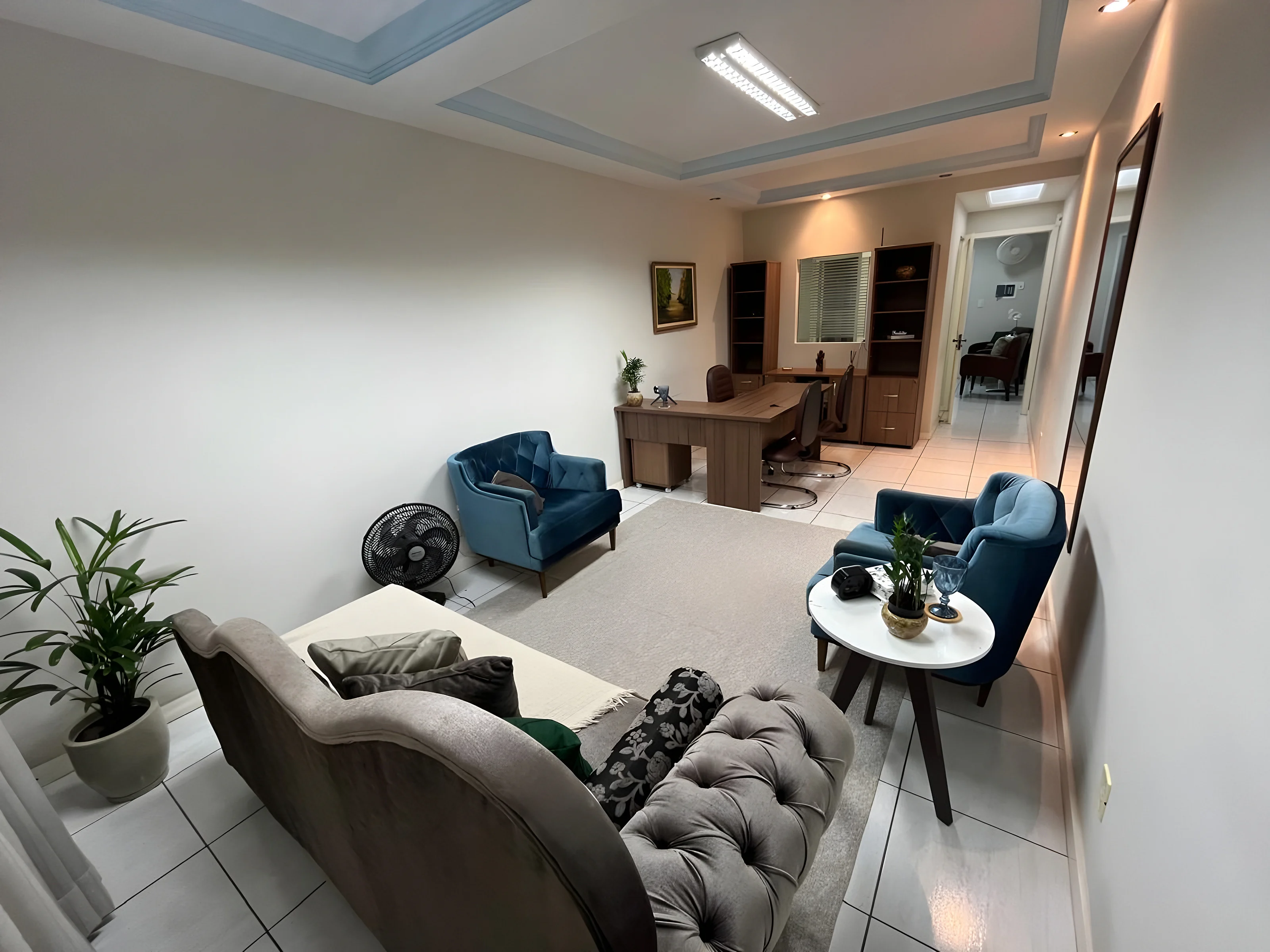 Sala de atendimento com sofá capitonê cinza, poltronas de veludo azul-petróleo e plantas